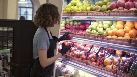 Close up of pretty caucasian worker in black apron and gloves stocking the fruits in supermarket. Young employee in glasses at work. Curly female arranging big red apples on shelf. Slow motion