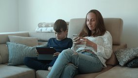 Mother  using her phone and her son watching on tablet at home and sitting on sofa
