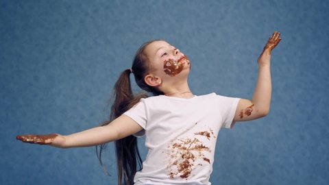 Cheerful little girl licking chocolate from her fingers. Dirty face and white t-shirt of a happy child. Funny kid in smeared chocolate isolated on blue.