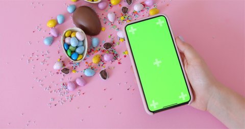 Female hand Holding Phone With Green Screen, iphone Mock-Up. Easter, junk-food, confectionery and unhealthy eating concept - chocolate eggs. Pink Background. 