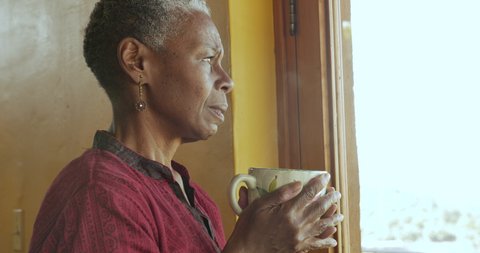 Concerned worried mature African American black woman holding a hot steaming tea or coffee cup looking out a window