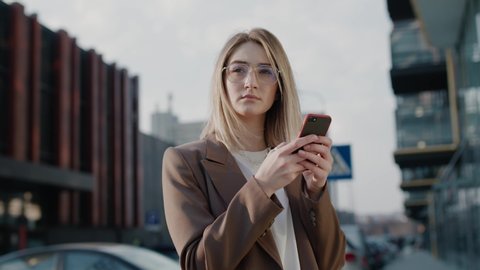 Portrait of blonde stylish caucasian young successful woman in glasses staying in the city center using phone texting message tapping scrolling professional female spring sun shine technology