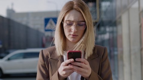Beautiful young serious businesswoman in glasses using modern smart phone while walking at break in the city, professional female employer typing text message on cellphone outside spring slow motion