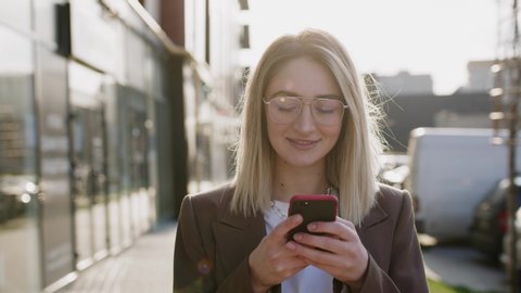 Stylish beautiful young caucasian woman in sunlight using phone, walking in the city center, smiling. Internet, business, outside, technology, eye, spring, mobile, portrait. Close up, slow motion.