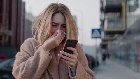 Caucasian young woman stand use phone feel sick coughs at outdoor fever cold allergy city disease female nose sneeze smartphone runny illness influenza cough district pneumonia covid19 slow motion