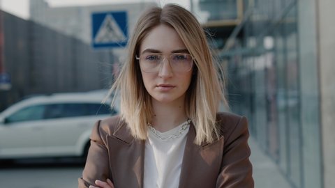 Portrait of stylish successful young businesswoman in formal jacket and glasses crossing her arms looking to the camera in the city center sunny day wind blowing her hair spring slow motion