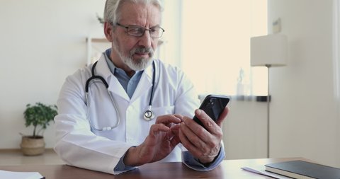 Happy elder senior male doctor wears white coat using smartphone app at workplace. Cheerful old adult surgeon or physician laughing watching funny video on cell, texting sms message in medical office.