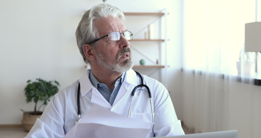 Concentrated serious old male doctor wearing white coat, stethoscope holding medical papers. Professional senior mature healthcare expert reading documents thinking of prescription, analyzing report. Royalty-Free Stock Footage #1048920529