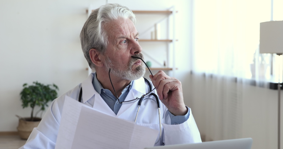 Concentrated serious old male doctor wearing white coat, stethoscope holding medical papers. Professional senior mature healthcare expert reading documents thinking of prescription, analyzing report. Royalty-Free Stock Footage #1048920529