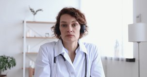Female doctor wears headset speaking looking at web cam providing remote medical assistance in virtual chat. Medic worker video calling consulting patient online. Telemedicine concept. Webcam view