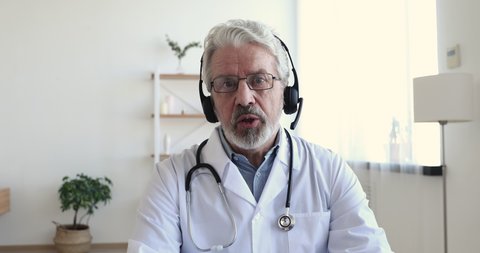 Senior male doctor wears white medical coat, stethoscope, headset makes distant video call. Old adult physician talks to camera consulting patient online in web chat. Telemedicine concept. Webcam view