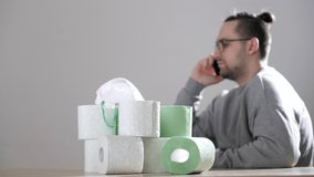 Handsome man sits next to stack of toilet paper rolls. Coronavirus or COVID-19. Pandemic worldwide, panic. Man holds smartphone and talking on the phone.