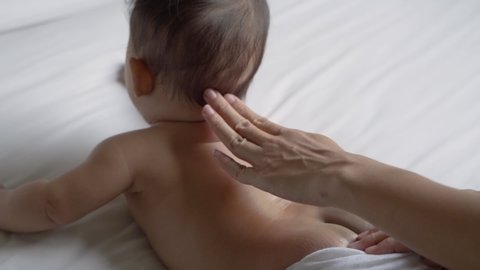 prone baby is oiled on his back by his mother while lying in the bedroom