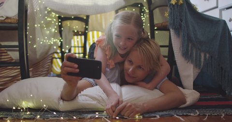 Happy Caucasian woman enjoying family time with her daughter at home together, smiling and talking in a tent in a sitting room, using a smartphone, taking selfie with her daughter, social distancing