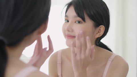 Beautiful young asian woman smiling look at mirror of checking face and skin care cream and cosmetic for rejuvenation and hygiene, beauty girl happy clean facial with lotion and for health.