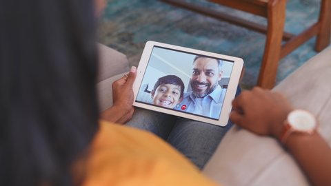 Rear view of young woman relaxing on couch while talking to her husband and son using digital tablet at home. Mature middle eastern man and child communicate through video chat on laptop. Happy family