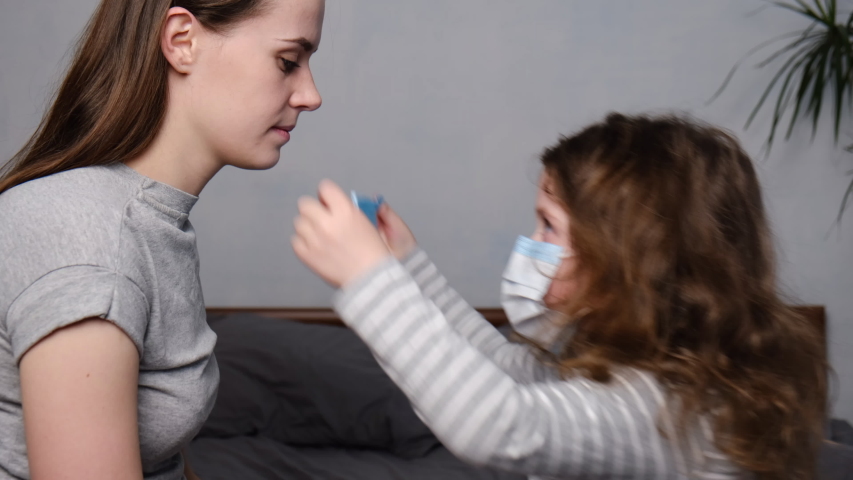 Cute daughter helping mother dressed medical mask sitting on bed, trying to protect family from contagious diseases. Concept of coronavirus or COVID-19 pandemic disease symptoms. Home quarantine. Royalty-Free Stock Footage #1048964155