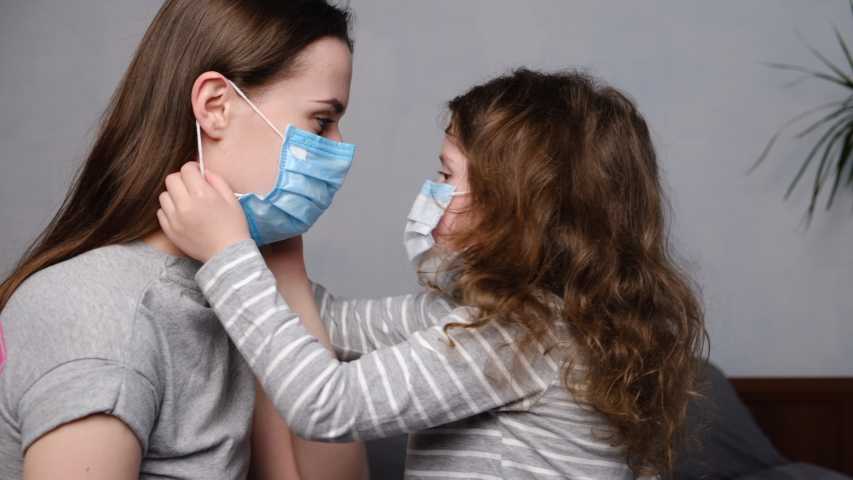 Cute daughter helping mother dressed medical mask sitting on bed, trying to protect family from contagious diseases. Concept of coronavirus or COVID-19 pandemic disease symptoms. Home quarantine. | Shutterstock HD Video #1048964155