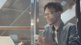 Young African American man putting in wireless earphones, waving at front camera of laptop, smiling and chatting with somebody via video call during workday in office