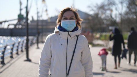 View of a stylish blonde woman in a white jacket and a protective mask against coronavirus going on a quay with lampposts on sunny day in spring in slo-mo
