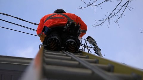 A telecoms operative is seen working from a ladder on a utility pole, wearing high visibility personal protective clothing, high viz PPE, and hard hat