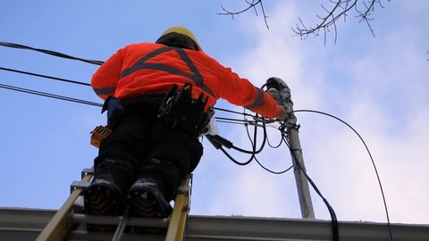 A telecoms operative man is seen working from a ladder on a utility pole, wearing high visibility personal protective clothing, high viz PPE, and hard hat