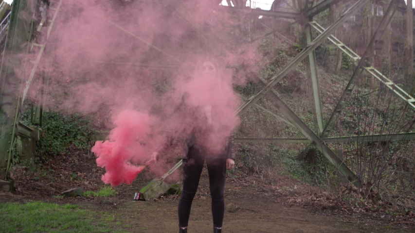 A young woman in a leather jacket waves around a pink smoke bomb under a bridge