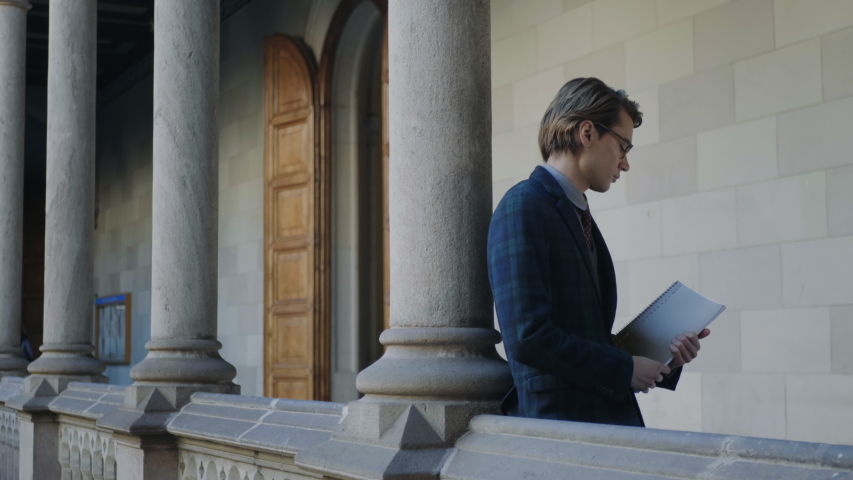 Portrait of young college student holding notebook in outdoor hallway. Handsome guy standing in campus building. Friendly businessman waving hand to colleague at outdoor meeting