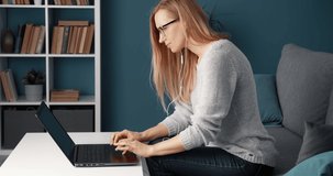 Side view of competent female worker in eyeglasses and casual outfit using laptop while sitting at couch. Mature woman with blond hair working at home.