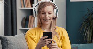 Happy blonde female in yellow sweater sitting on grey sofa with wireless headphones and holding smartphone. Mature woman searching favorite song from playlist.
