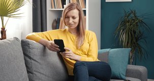 Pretty blonde lady sitting at living room with modern design and surfing internet on smartphone. Mature woman in casual outfit relaxing at home.