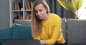 Mature lady with blond hair looking at computer screen and showing real emotions about successful business project. Happy woman in yellow sweater enjoying remote work at home.