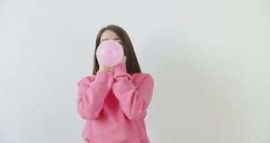 Young woman blowing balloon over white wall background . Girl in pink hoodie having fun at home or studio. 4k raw video footage slow motion