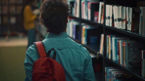 University Library: Student Walks Between Rows of Bookshelves Searching for the Right Book Title for Assignment and Exam Preparations. Back View Following Slow Motion Camera Shot. Young People Study