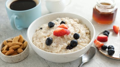 Eating healthy breakfast food. Add fresh summer berries to oatmeal porridge. Breakfast table with cup of black coffee, honey, bowl of oatmeal and berries