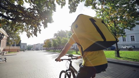 The city courier service, the courier in a yellow T-shirt with a yellow thermal backpack rides a bicycle, the video shot is from the back.