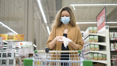 A woman in a medical mask puts on protective rubber gloves to protect against coronavirus in a supermarket, takes a grocery cart and goes to get food. Protective measures against the spread of the