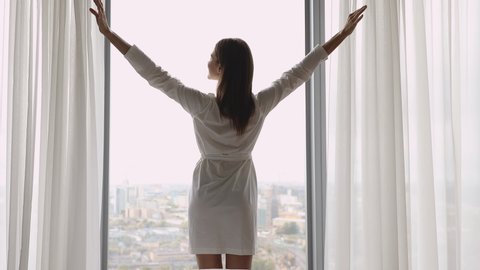Rear view happy young brunette woman in white silk nightgown standing near huge panoramic window with opened light curtains, raising arms, stretching back, welcoming new day, good morning time.