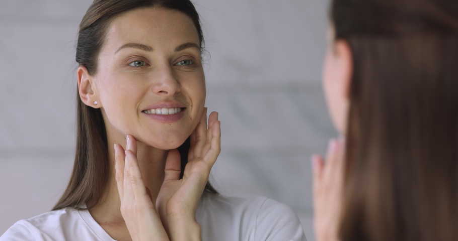 Close up head shot young well-groomed lady looking at mirror in bathroom, touching face, feeling satisfied with perfect healthy skin condition after professional cosmetics, moisturizing treatment. Royalty-Free Stock Footage #1049015203