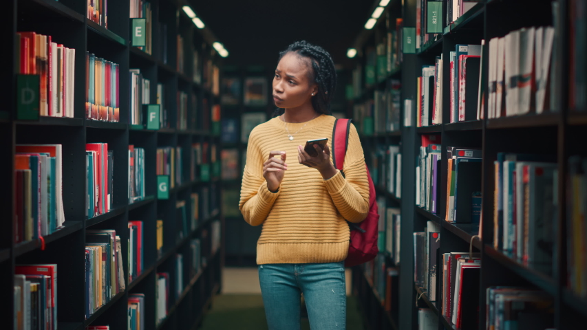 University Library: Portrait of Gifted Beautiful Black Girl Walking Between Rows of Bookshelves Using Smartphone Searching for the Right Book Title, Finds and Picks one for Class Assignment Royalty-Free Stock Footage #1049020678