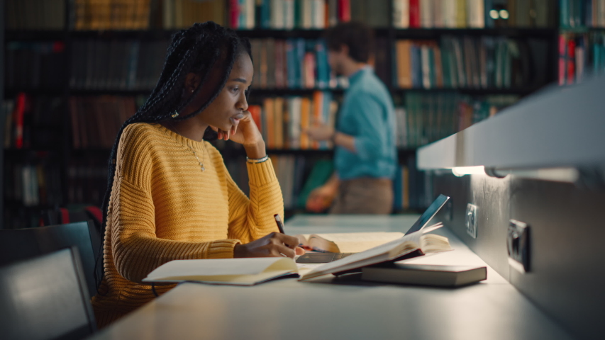 University Library: Gifted Black Girl uses Laptop, Writes Notes for the Paper, Essay, Study for Class Assignment. Students Learning, Studying for Exams College. Side View Portrait with Bookshelves Royalty-Free Stock Footage #1049020681