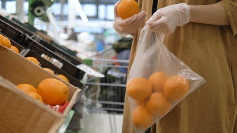 A girl in a supermarket selects fresh oranges in rubber gloves and puts them in a plastic bag. Healthy nutrition to increase immunity. Protective measures against the coronavirus pandemic.