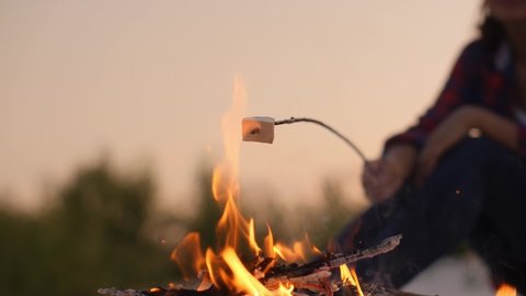 Close up photo of roasting food over the fire near tent in camping. Focus on fire. Young tourist woman having fun near the fire in the nature.