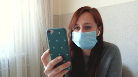 Woman in a medical protective face mask at home quarantine. Girl using mobile phone, video call. Pandemic mood. Woman speaking online, internet communication. Self-isolation during epidemic.