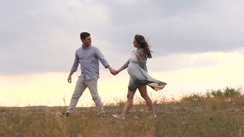 Happy young man leads his pregnant wife by the hand in the field with dry grass at sunset. Happy pregnant family walking in the field outside the city. Follow me.