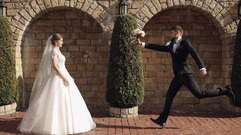 Newlyweds. Lovely young caucasian bride and groom in the park near stone wall. Groom jumps to bride. Wedding couple. Happy family. Man and woman in love. Slow motion