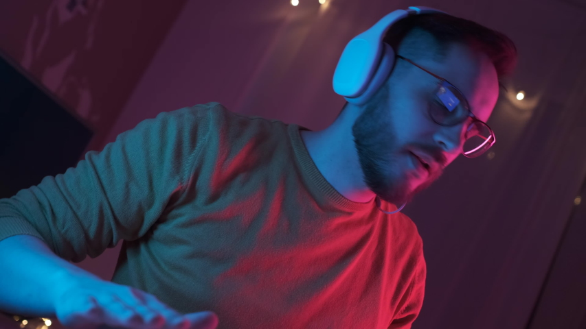 Portrait young handsome man in headphones listening energetic music at home. Bearded student with glasses relax in living room in colourful neon light. Dance hand movements to the rhythm. Indoors Royalty-Free Stock Footage #1049063671