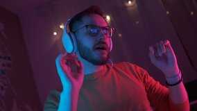 Portrait young handsome man in headphones listening energetic music at home. Bearded student with glasses relax in living room in colourful neon light. Dance hand movements to the rhythm. Indoors - Powered by Shutterstock - Get 15% off with code: PIKWIZARD15
