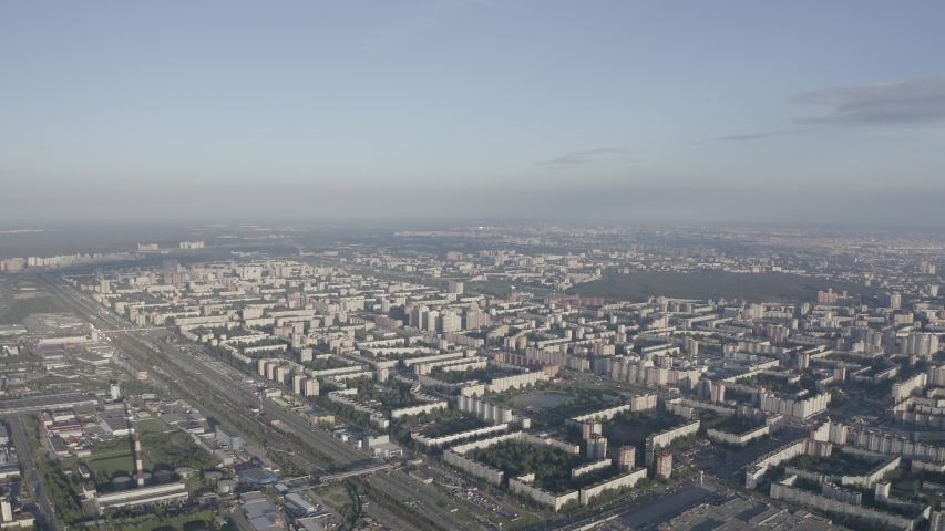 Panoramic view of St. Petersburg from the air during sunset. A sweeping aerial cityscape reveals a vast residential district of long rectangular apartment blocks, pale concrete facades, and enclosed green courtyards spreading across a flat urban plain. Multilane highways cut between the housing estates, while patches of parkland and a wide river enter the right side of the view. The camera glides forward at a steady altitude, keeping the horizon level as foreground blocks pass beneath and the city stretches toward a hazy distance. A slight rightward drift gradually brings the water and greener outskirts into the frame, while traffic and rooflines remain mostly still within the vast grid. Soft sunset light washes the scene in muted gray, beige, and blue tones, with thin clouds hanging above a pale sky. The atmosphere is calm and expansive, capturing the scale and repetition of the sleeping areas on the edge of the metropolis. D-Log.