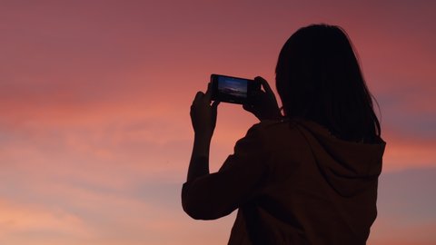 Back view of Asian woman hand holding smartphone Take Photo on the sunset at the camping beach of Thailand. Evening sky scene with golden light from the setting sun in summer